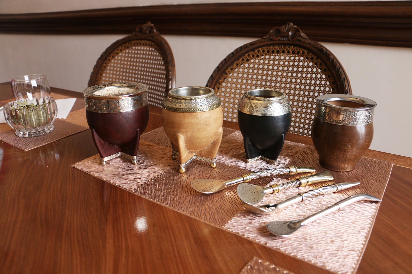 Collection of various traditional leather-wrapped mate cups with decorative silver rims, accompanied by several ornate metal bombillas on a wooden table