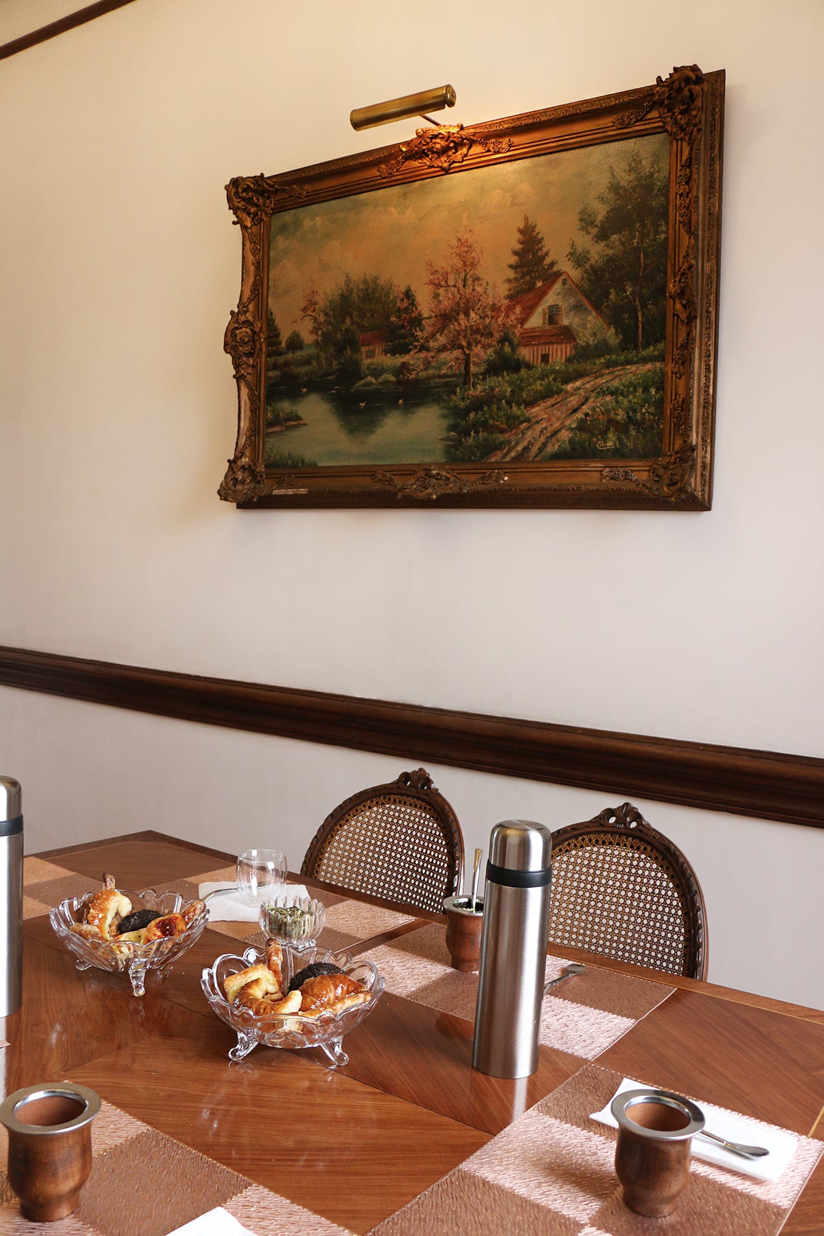 View of a classic dining room with a traditional painting on the wall, featuring a wooden table set for a mate tasting with mate cups, thermoses, and glass bowls