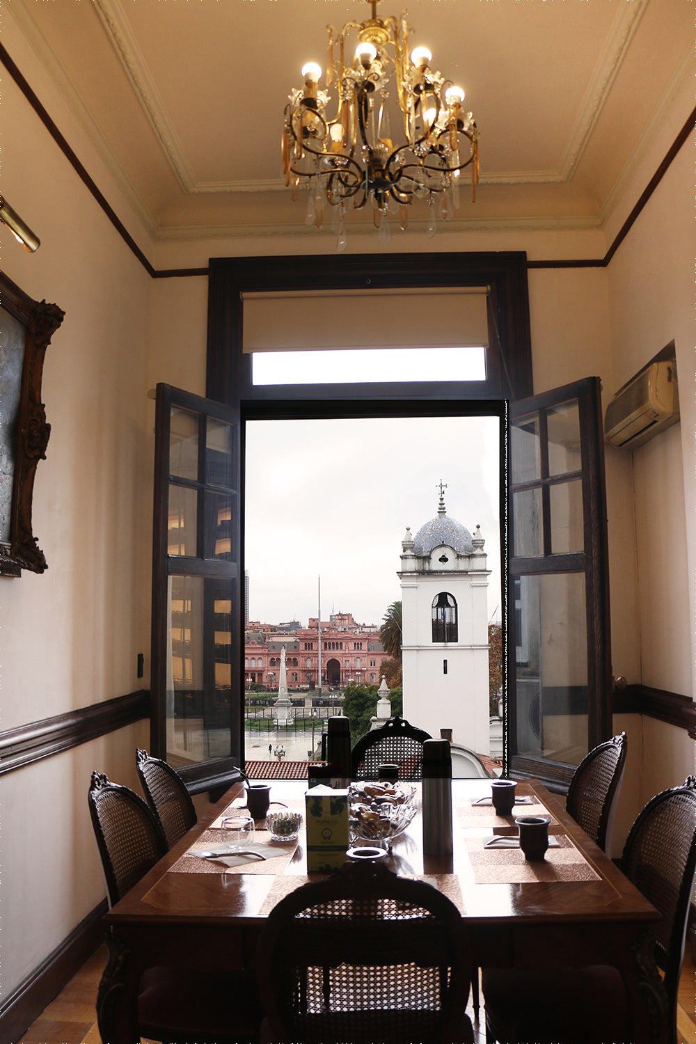 An interior view of a historic Argentine room with a dining set and chandelier, featuring a stunning view of Plaza de Mayo and the Cathedral