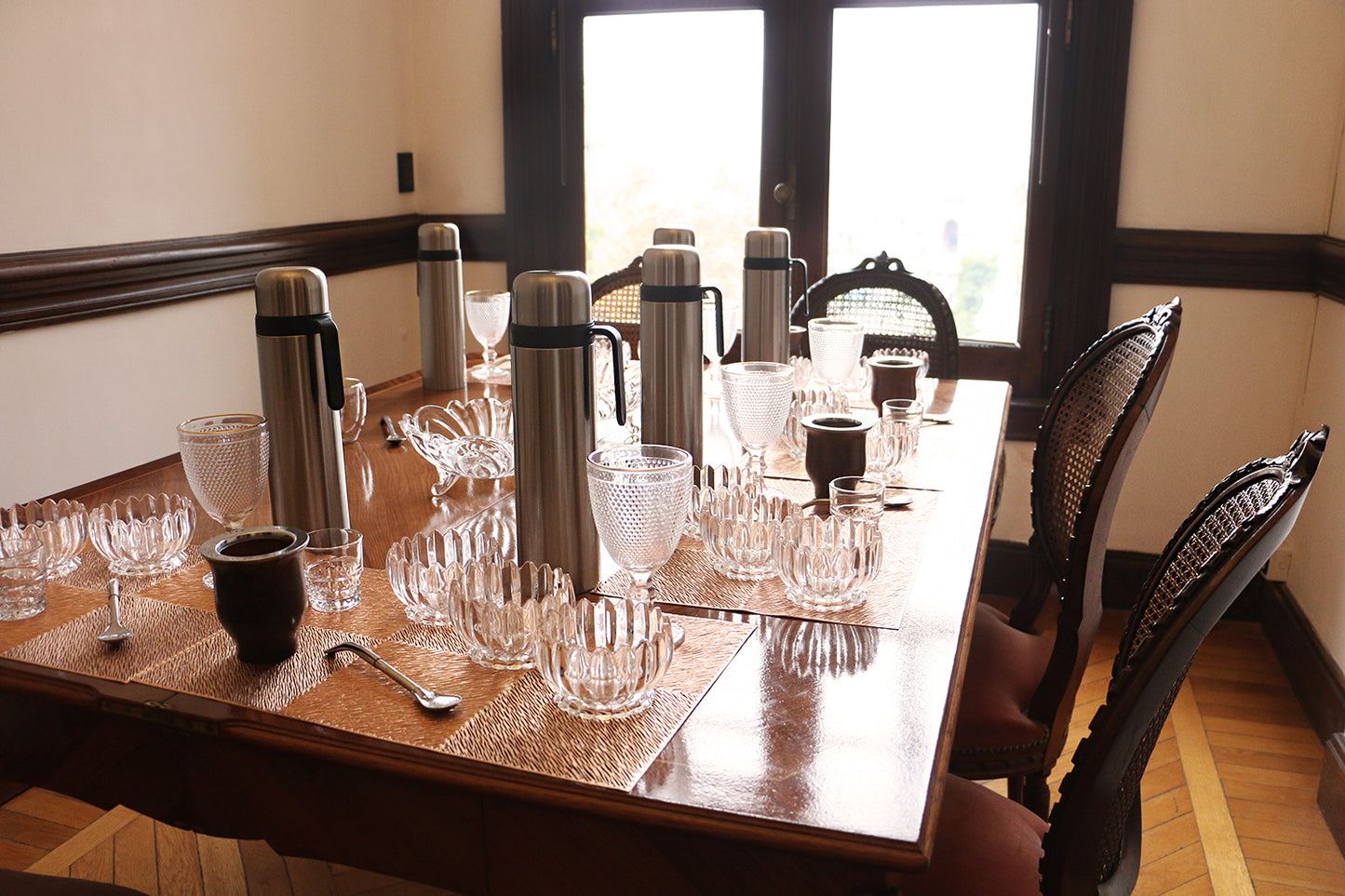 Detailed view of a table set for a mate tasting, featuring various glass cups, metal thermoses, and mate gourds on a wooden table
