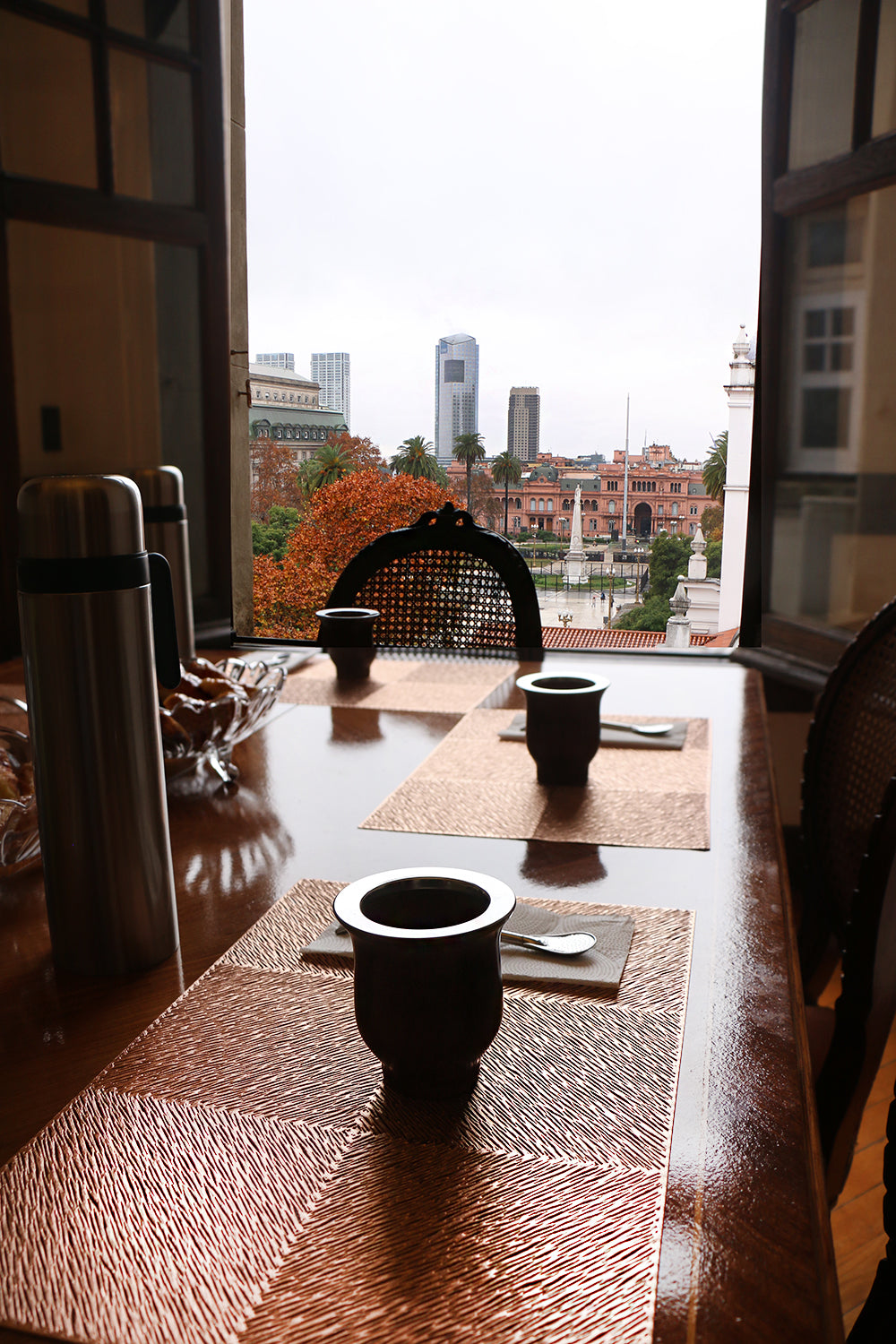 View from inside a room of a wooden table set for a mate tasting, overlooking the Plaza de Mayo and surrounding cityscape in Buenos Aires