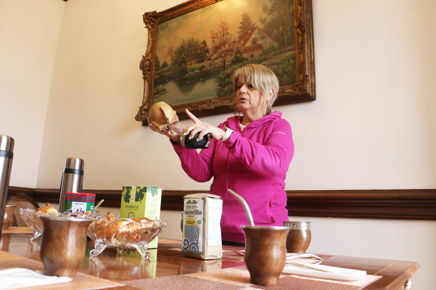 Woman demonstrating and explaining the preparation of traditional mate to a group, holding a gourd with yerba mate in a setting with various mate accessories
