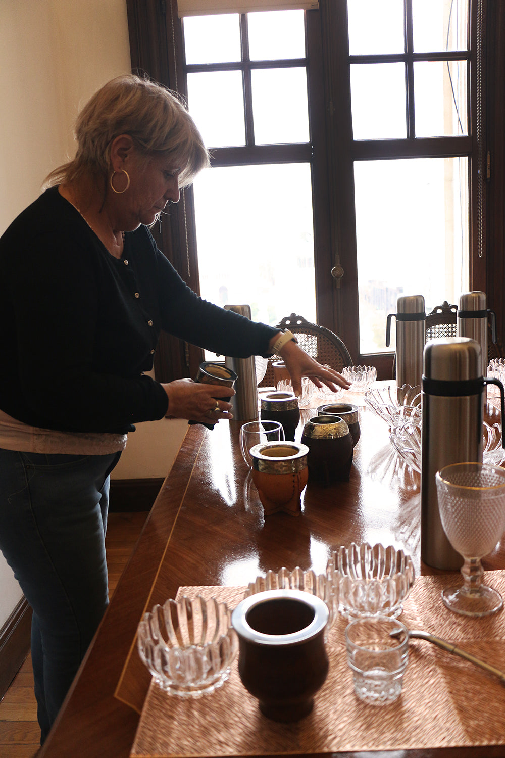 Woman arranging various traditional mate cups and glass bowls on a wooden table for a mate tasting session