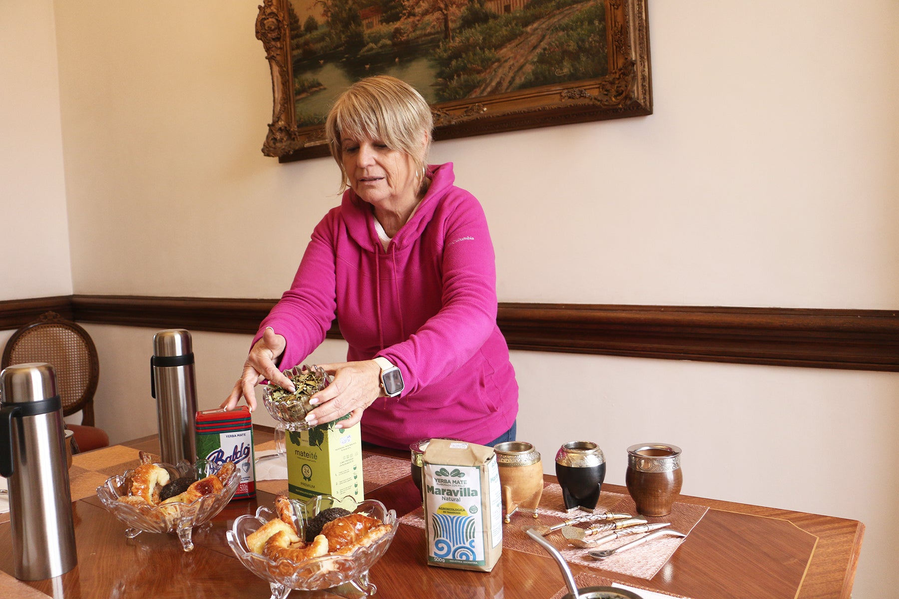 Woman pouring yerba mate from a small bowl into a container, with various mate cups and packages on a wooden table for a tasting session